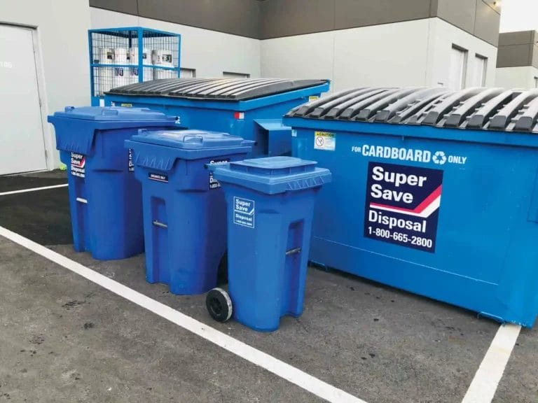 A row of blue recycling bins labelled "Super Save Disposal" with two large dumpsters for cardboard only, situated in a parking area near a building
