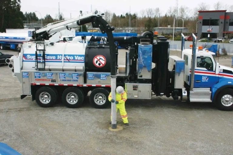 A worker in high-visibility gear operates the hose of a Super Save Hydro Vac truck, performing excavation work on a gravel surface