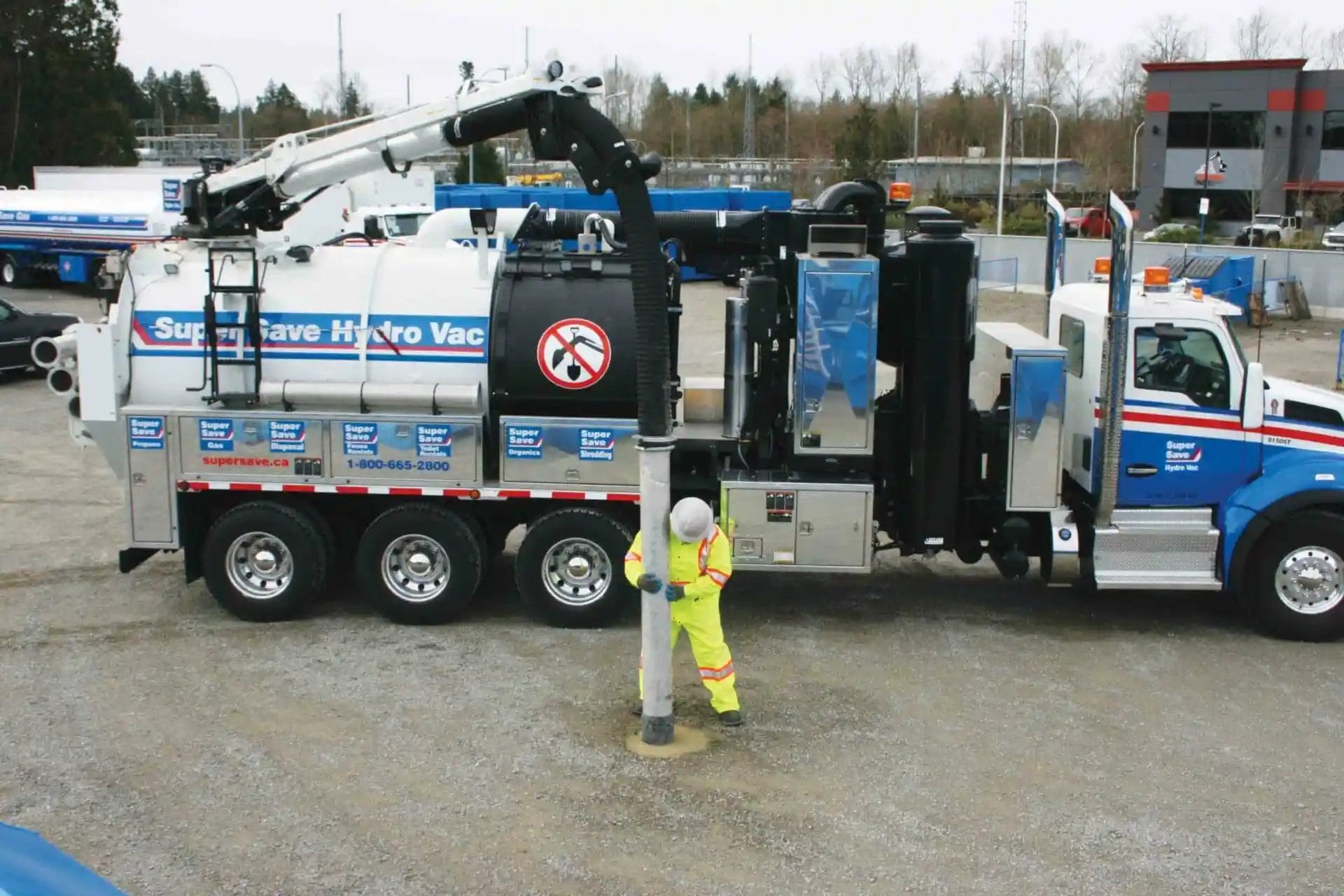 A worker in high-visibility gear operates the hose of a Super Save Hydro Vac truck, performing excavation work on a gravel surface