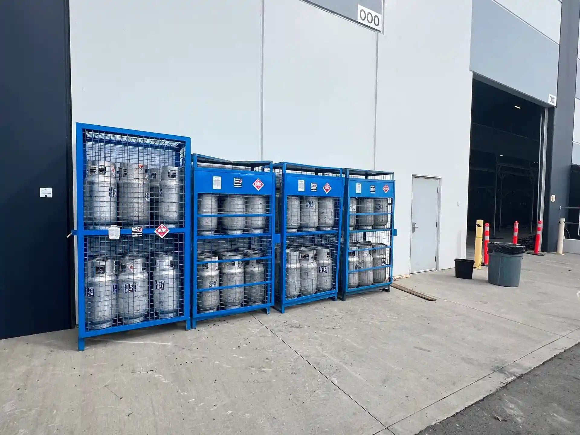 Storage area with blue metal cages containing several gas cylinders, labeled with safety warning signs, positioned outside a warehouse. A door and a garbage bin are visible in the background.