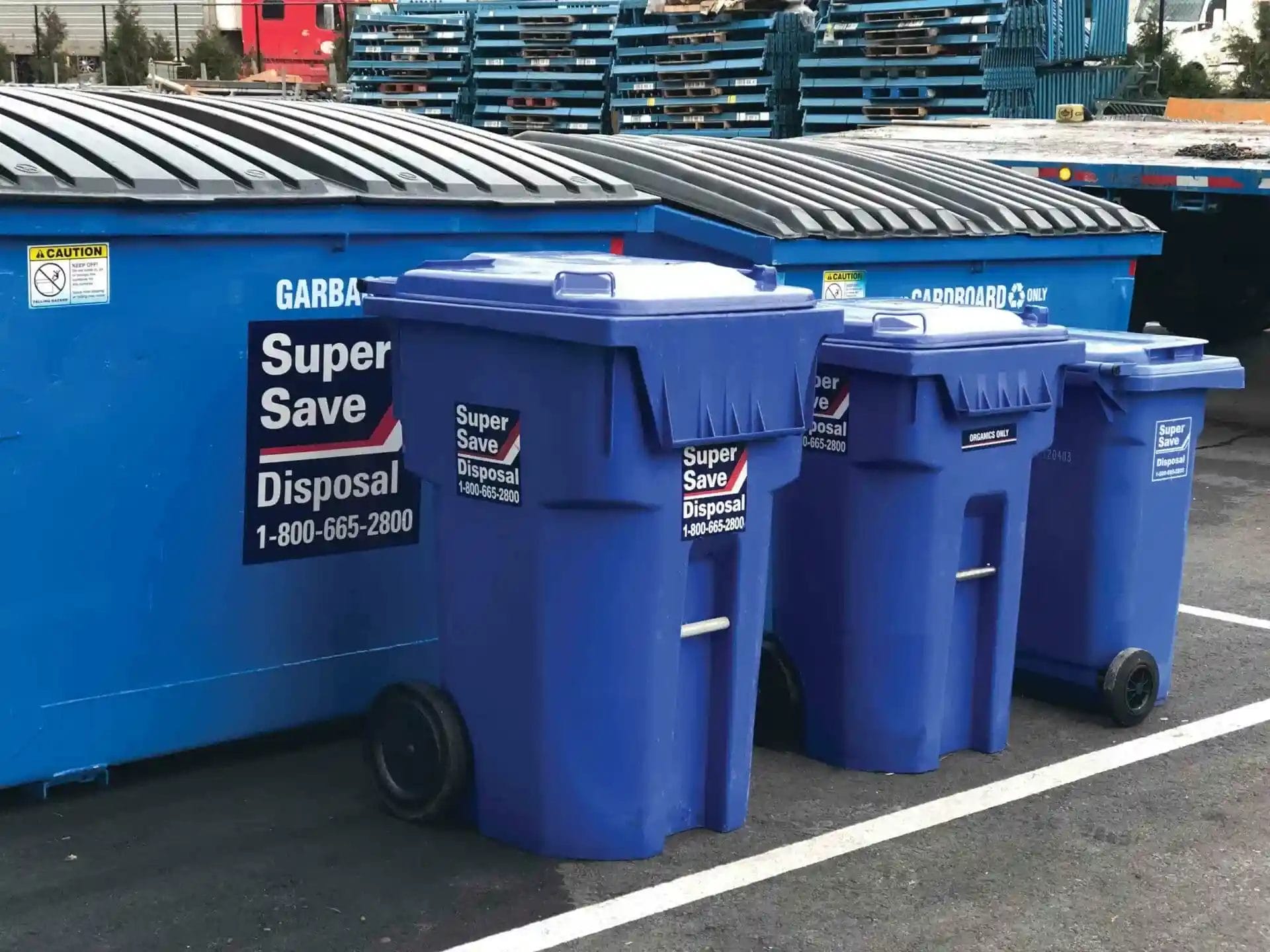 Three blue waste disposal bins labeled "Super Save Disposal" are positioned beside a larger blue dumpster in a parking lot. Pallets are stacked in the background