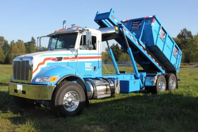 A large blue and white roll-off bin truck in a grassy field with its rear container lifted. The vehicle features "Super Save Disposal" branding and is situated in front of trees.
