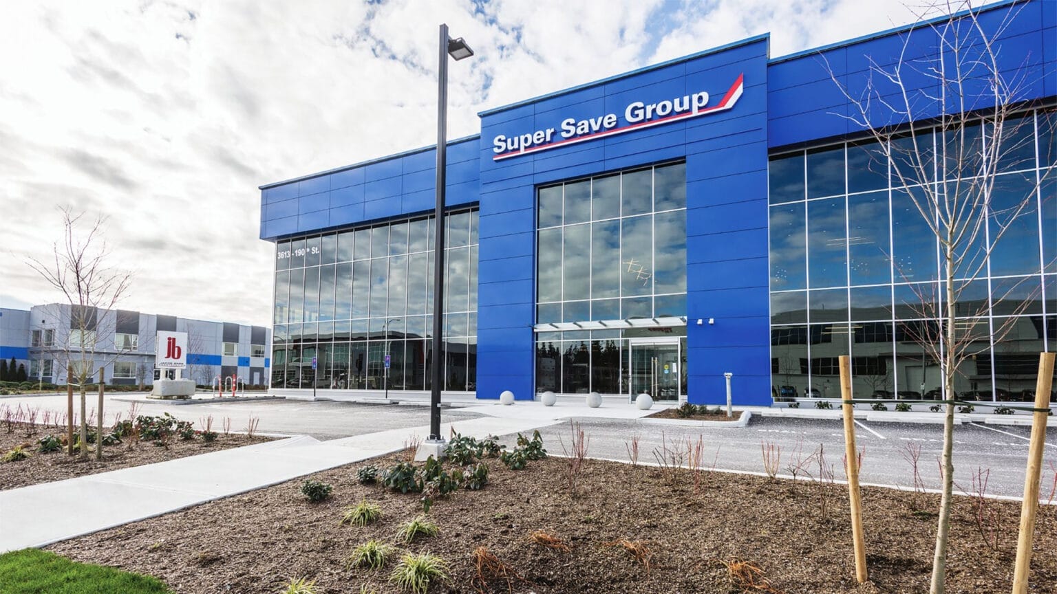 Exterior view of a modern building with glass windows and a blue facade, labelled "Super Save Group." The surrounding area features a landscaped garden with small plants and a rock bed