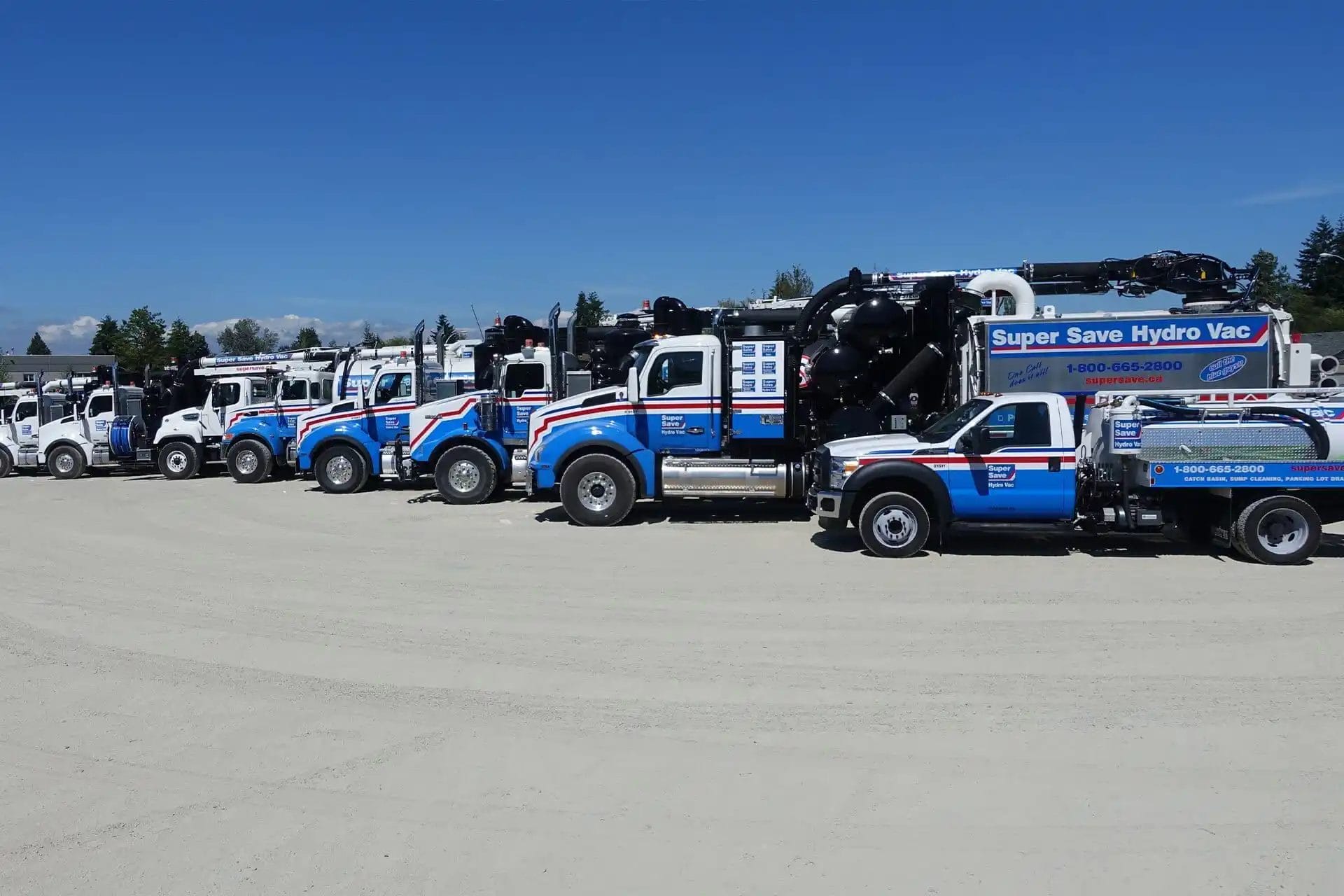 A line of Super Save Hydro Vac trucks parked on a sunny day in a dirt lot with a clear blue sky. The trucks are painted in blue, white, and red with company branding.
