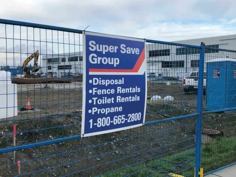 A construction site with a blue fence displaying a sign for "Super Save Group" offering disposal, fence rentals, toilet rentals, and propane services
