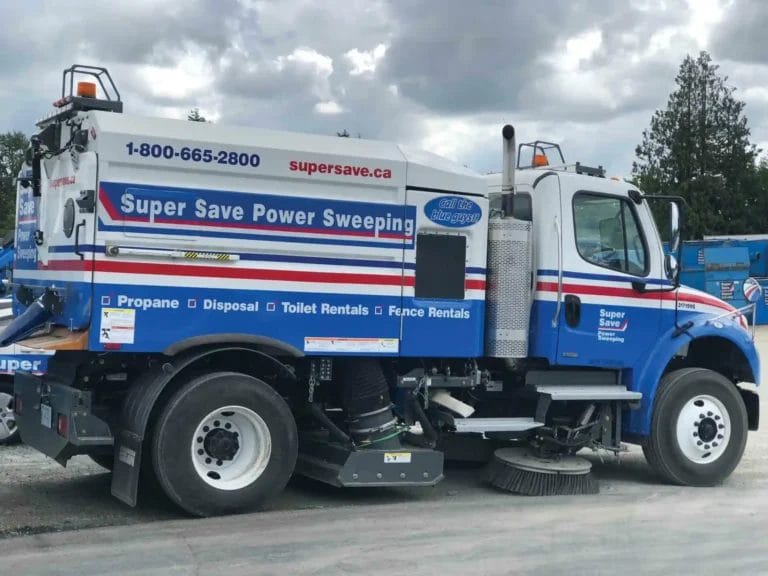 Side view of a blue and white street sweeper truck with "Super Save Power Sweeping" and contact information on the side, parked on a gravel area with trees and a cloudy sky in the background