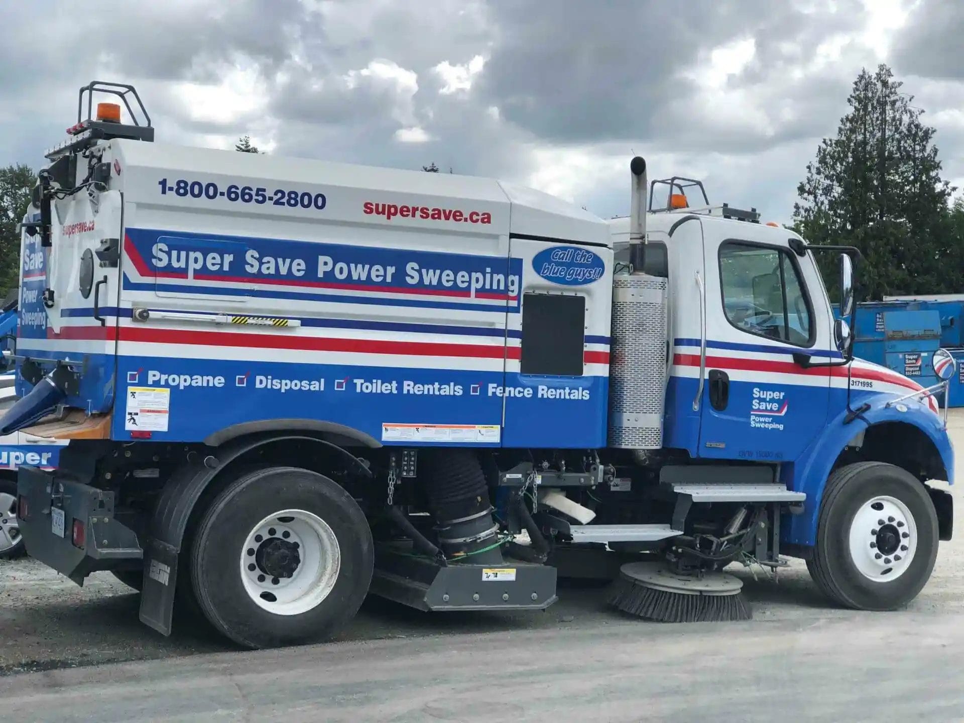 Side view of a blue and white street sweeper truck with "Super Save Power Sweeping" and contact information on the side, parked on a gravel area with trees and a cloudy sky in the background